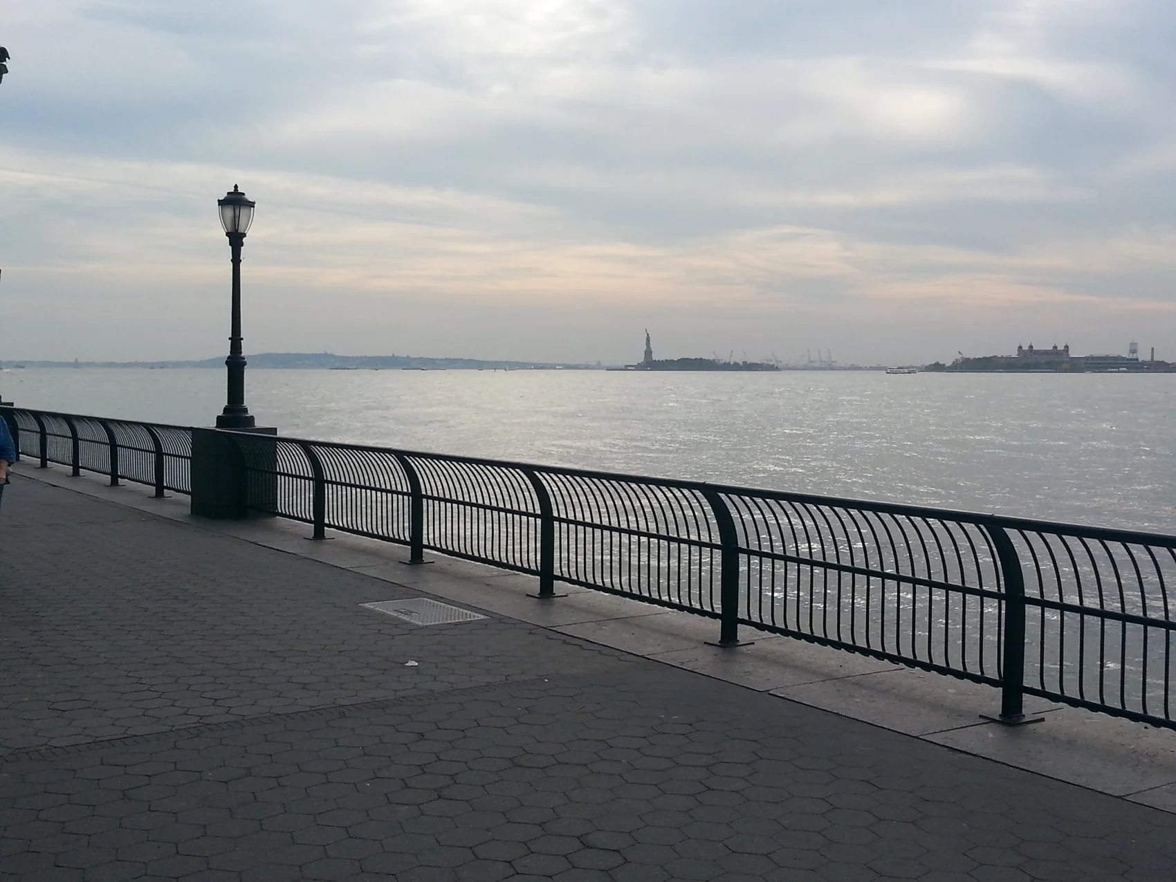 Waterfront promenade with a streetlight and a view across the water toward the Statue of Liberty